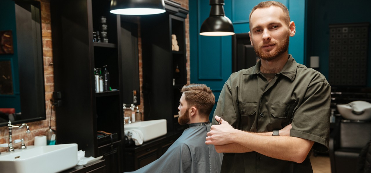 happy-young-hairdresser-standing-near-his-client-looking-at-camera-SBI-302894619